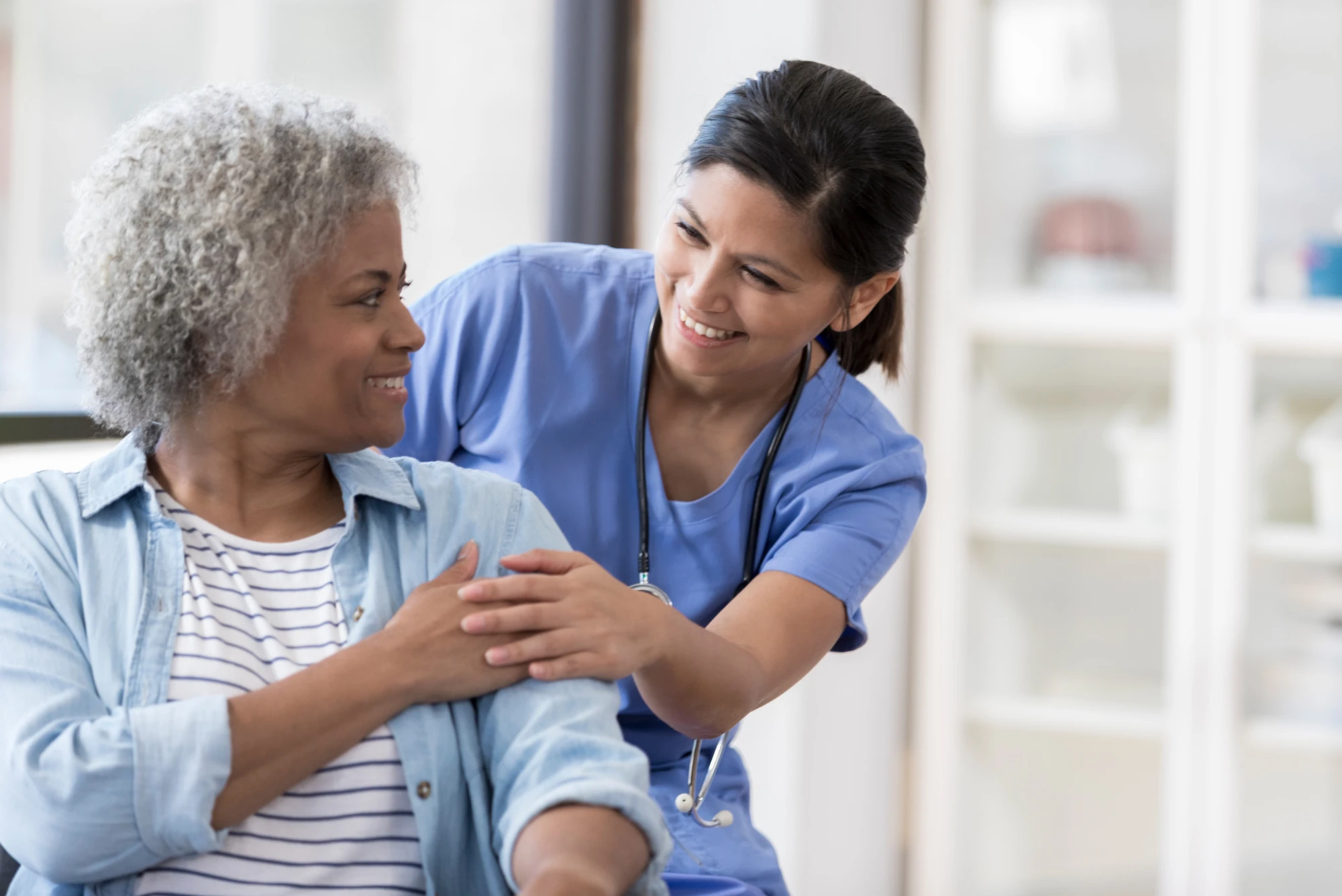 A nurse in blue scrubs rests a hand on an older patient's arm during an unhurried exam room visit.