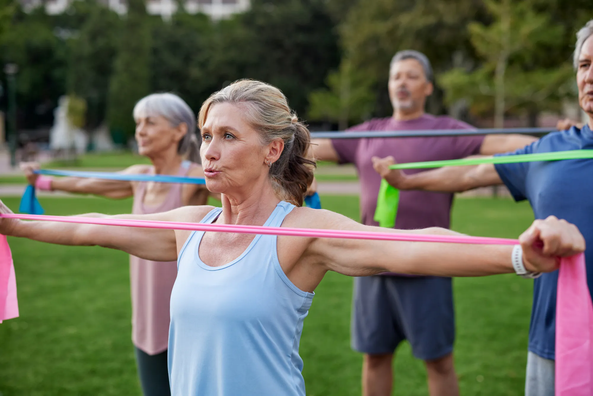 A group of older adults in an outdoor fitness class stretch resistance bands across their chests, illustrating the load-based and resistance training central to protecting the bone-muscle unit.