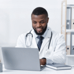 Smiling doctor in a white coat sitting at a desk and working on a laptop.