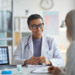 Female physician with glasses speaking with a patient at her desk with brain scans visible.